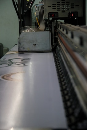 A large industrial machine standing in a factory environment is captured up-close. The machine appears to be a printer or fabric printing device. The conveyor belt on the machine has a visible pattern, and there are various mechanical parts visible, showing its operational complexities.