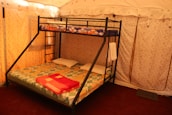 A cozy interior features a bunk bed with a double mattress on the bottom and a single mattress on top, each with colorful patterned bedspreads. The bed frame is made of black metal with a ladder for the upper bunk. The room has beige fabric walls and ceiling, with soft lighting creating a warm atmosphere. A folded red blanket and two white pillows are placed on the lower bed, while a towel hangs on the frame.