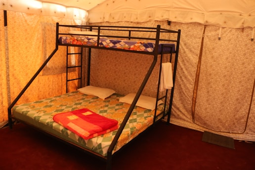 A cozy interior features a bunk bed with a double mattress on the bottom and a single mattress on top, each with colorful patterned bedspreads. The bed frame is made of black metal with a ladder for the upper bunk. The room has beige fabric walls and ceiling, with soft lighting creating a warm atmosphere. A folded red blanket and two white pillows are placed on the lower bed, while a towel hangs on the frame.