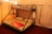 A cozy interior features a bunk bed with a double mattress on the bottom and a single mattress on top, each with colorful patterned bedspreads. The bed frame is made of black metal with a ladder for the upper bunk. The room has beige fabric walls and ceiling, with soft lighting creating a warm atmosphere. A folded red blanket and two white pillows are placed on the lower bed, while a towel hangs on the frame.
