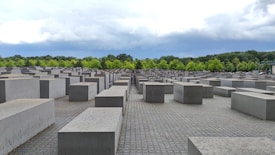 A vast array of large concrete rectangular blocks arranged in a grid pattern across a paved area. The sky is overcast with thick clouds, and a line of lush green trees can be seen in the background, adding contrast to the gray blocks.
