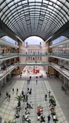 An indoor shopping mall with a high arched glass ceiling allowing natural light to illuminate the space. The two floors of shops have glass railings and are lined with various stores. Below, people walk and socialize near small tables with chairs and potted plants, creating a lively atmosphere.