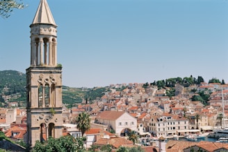 A picturesque coastal town featuring a tall, historic stone tower with arches and intricate carvings. The foreground displays lush greenery and palm trees, while the background reveals a dense arrangement of charming, red-tiled rooftops and stone buildings nestled against a backdrop of rolling hills and a clear blue sky. Boats are visible docked at the waterfront.