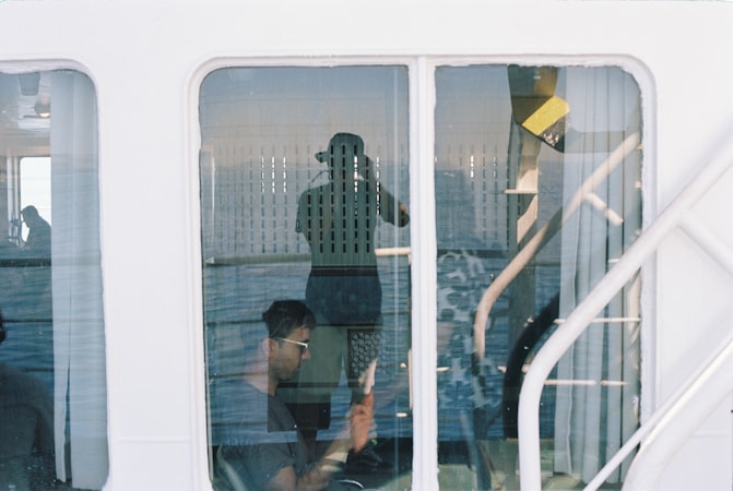 A reflection in a window shows a person taking a photo with a camera, along with a second person sitting inside, possibly using a smartphone or other device. The interior appears to be part of a vehicle or vessel, suggested by the white metal framework and visible water, hinting at a journey or transit.
