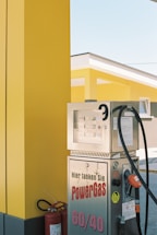 A brightly colored gas station featuring a yellow and black color scheme. Several metallic gas pumps are visible, with the text 'Hier tanken Sie PowerGas 60/40' printed on one of them. A fire extinguisher is placed nearby, and the modern architecture of the station is highlighted in the background.