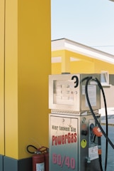 A brightly colored gas station featuring a yellow and black color scheme. Several metallic gas pumps are visible, with the text 'Hier tanken Sie PowerGas 60/40' printed on one of them. A fire extinguisher is placed nearby, and the modern architecture of the station is highlighted in the background.