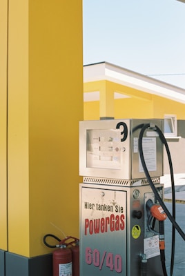 A brightly colored gas station featuring a yellow and black color scheme. Several metallic gas pumps are visible, with the text 'Hier tanken Sie PowerGas 60/40' printed on one of them. A fire extinguisher is placed nearby, and the modern architecture of the station is highlighted in the background.