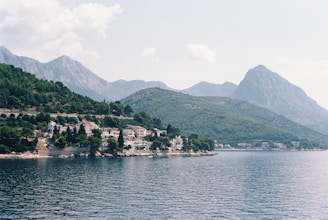A scenic coastline featuring a series of residential buildings nestled among dense greenery. Mountains rise majestically in the background under a partly cloudy sky. The calm water of the ocean stretches out in the foreground, reflecting the serene atmosphere.