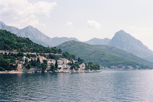 A scenic coastline featuring a series of residential buildings nestled among dense greenery. Mountains rise majestically in the background under a partly cloudy sky. The calm water of the ocean stretches out in the foreground, reflecting the serene atmosphere.