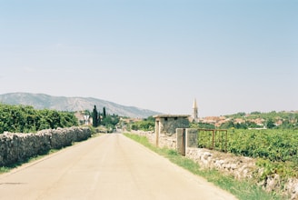 A rural road stretches into the distance, flanked by stone walls and lush greenery. A small village with a prominent church tower is visible in the middle ground. Rolling hills create a scenic backdrop under a clear blue sky.
