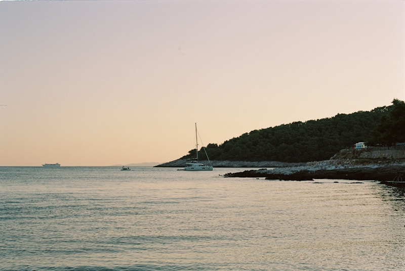 A serene coastal scene at sunset with a sailboat anchored near a rocky shoreline. The sky is a soft gradient of pastel colors, and the sea reflects these tones. In the distance, a larger ship is visible on the horizon, and a car is parked near the trees lining the shore.
