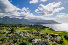 A scenic view of a golf course surrounded by residential houses with lush greenery, set against a backdrop of mountains and the ocean. The sky is partly cloudy, casting shadows over the landscape.