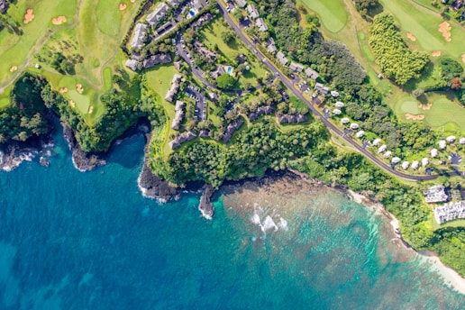 An aerial view of a coastal area featuring lush greenery, residential buildings, and a winding road. The landscape includes a golf course with neatly trimmed grass and sand bunkers. The vibrant blue ocean meets the rocky shoreline, creating a picturesque contrast.