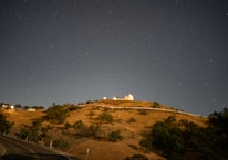 A hilltop observatory illuminated against a clear night sky filled with stars. The landscape includes grassy slopes, scattered trees, and a winding road leading up the hill. The observatory dome is a prominent feature, with its white structure and surrounding buildings visible.