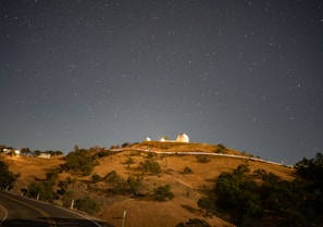 A hilltop observatory illuminated against a clear night sky filled with stars. The landscape includes grassy slopes, scattered trees, and a winding road leading up the hill. The observatory dome is a prominent feature, with its white structure and surrounding buildings visible.