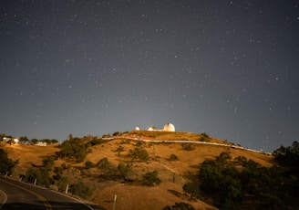 A hilltop observatory illuminated against a clear night sky filled with stars. The landscape includes grassy slopes, scattered trees, and a winding road leading up the hill. The observatory dome is a prominent feature, with its white structure and surrounding buildings visible.