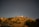 A hilltop observatory illuminated against a clear night sky filled with stars. The landscape includes grassy slopes, scattered trees, and a winding road leading up the hill. The observatory dome is a prominent feature, with its white structure and surrounding buildings visible.
