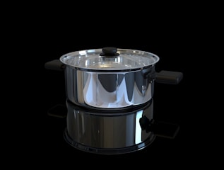 A shiny stainless steel pot with a glass lid is positioned against a black background. The pot has black handles on either side and a matching black knob on the lid. It is highly reflective, capturing light and mirrored reflections of its surroundings.