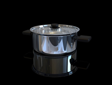 A shiny stainless steel pot with a glass lid is positioned against a black background. The pot has black handles on either side and a matching black knob on the lid. It is highly reflective, capturing light and mirrored reflections of its surroundings.