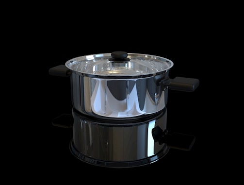 A shiny stainless steel pot with a glass lid is positioned against a black background. The pot has black handles on either side and a matching black knob on the lid. It is highly reflective, capturing light and mirrored reflections of its surroundings.