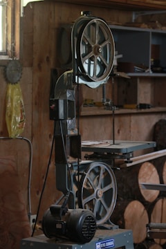 A workshop featuring a band saw machine with a large wheel and a cutting table. The workshop has wooden walls and shelves filled with tools and materials. There are circular wooden logs on the floor and various items scattered on the shelves in the background.