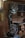 A workshop featuring a band saw machine with a large wheel and a cutting table. The workshop has wooden walls and shelves filled with tools and materials. There are circular wooden logs on the floor and various items scattered on the shelves in the background.