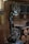 A workshop featuring a band saw machine with a large wheel and a cutting table. The workshop has wooden walls and shelves filled with tools and materials. There are circular wooden logs on the floor and various items scattered on the shelves in the background.