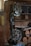A workshop featuring a band saw machine with a large wheel and a cutting table. The workshop has wooden walls and shelves filled with tools and materials. There are circular wooden logs on the floor and various items scattered on the shelves in the background.