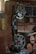 A workshop featuring a band saw machine with a large wheel and a cutting table. The workshop has wooden walls and shelves filled with tools and materials. There are circular wooden logs on the floor and various items scattered on the shelves in the background.
