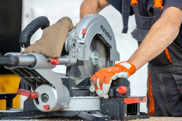 A person is operating a circular miter saw, likely cutting through a piece of wood. The individual's gloved hands are holding the saw steady, and the saw is branded with 'Metabo'. Sawdust is scattered around, suggesting active work.