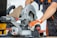 A person is operating a circular miter saw, likely cutting through a piece of wood. The individual's gloved hands are holding the saw steady, and the saw is branded with 'Metabo'. Sawdust is scattered around, suggesting active work.