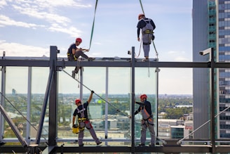 Construction workers in safety gear, including helmets and harnesses, are installing large glass panels on a high-rise building. The cityscape is visible in the background, providing a scenic view.