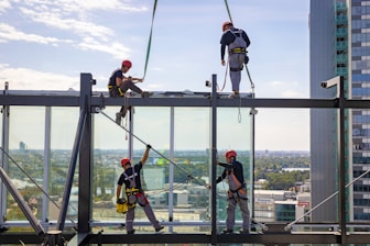 Construction workers in safety gear, including helmets and harnesses, are installing large glass panels on a high-rise building. The cityscape is visible in the background, providing a scenic view.