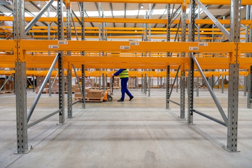 Inside a large warehouse with empty metal racks painted in bright yellow. A worker in a yellow and blue uniform is transporting a pallet with a manual pallet jack.