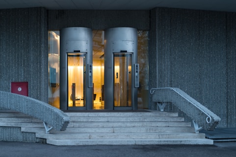 Two modern glass door elevators are illuminated with warm lighting, situated at the top of a short flight of stone steps. The steps are flanked by curved metal railings leading to the elevators. The building&rsquo;s exterior is covered in vertical concrete panels, adding a textured appearance. A red fire equipment box is visible on the wall to the left.