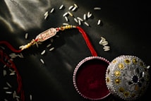 A decorative red thread bracelet with a golden centerpiece is placed on a dark surface. Scattered around it are white rice grains. Next to the bracelet, an intricately designed container with a beaded lid is open, revealing a deep red powder inside.