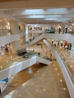 A spacious, multi-level shopping mall interior features several levels connected by escalators. Shoppers can be seen walking and browsing store fronts. The flooring is polished and the lighting is bright, highlighting the modern architectural design.