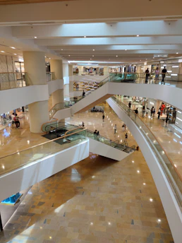 A spacious, multi-level shopping mall interior features several levels connected by escalators. Shoppers can be seen walking and browsing store fronts. The flooring is polished and the lighting is bright, highlighting the modern architectural design.