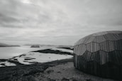 A geodesic dome made of wooden panels is set against a stark, arctic landscape. Snow patches cover the rocky terrain, leading to a calm body of water with distant, snow-capped hills under a cloudy sky.