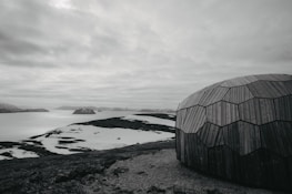 A geodesic dome made of wooden panels is set against a stark, arctic landscape. Snow patches cover the rocky terrain, leading to a calm body of water with distant, snow-capped hills under a cloudy sky.