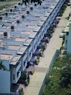 A row of modern, uniformly designed white residential buildings with flat roofs and balconies. The structures are aligned closely with a driveway separating them. The driveway has parked vehicles, including a visible white car and a bicycle. Surrounding the area are small patches of greenery, and a fence distinguishes the buildings from an adjacent grassy area.
