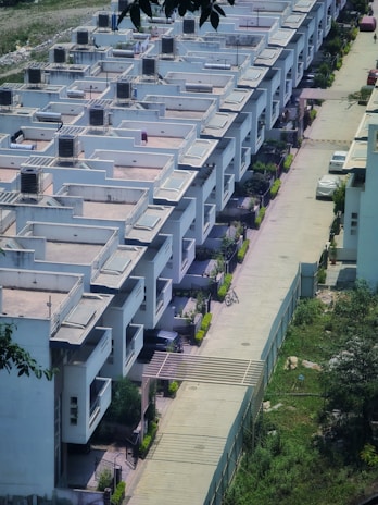 A row of modern, uniformly designed white residential buildings with flat roofs and balconies. The structures are aligned closely with a driveway separating them. The driveway has parked vehicles, including a visible white car and a bicycle. Surrounding the area are small patches of greenery, and a fence distinguishes the buildings from an adjacent grassy area.