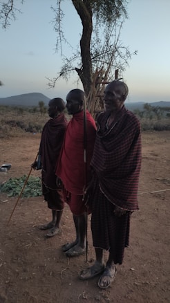 Three individuals are standing in the foreground, draped in traditional checkered garments. They are each holding long sticks, set against a background of dry, open plains with scattered trees and hills in the distance. A tree with sparse branches rises behind them.