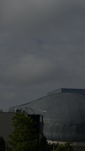 A large, dome-like building made of metal with a ribbed pattern is set against a cloudy sky. The structure appears industrial, with an attached rectangular section possibly made of concrete. Trees with green foliage are visible in the foreground, adding a natural contrast to the urban architecture.