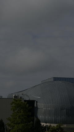 A large, dome-like building made of metal with a ribbed pattern is set against a cloudy sky. The structure appears industrial, with an attached rectangular section possibly made of concrete. Trees with green foliage are visible in the foreground, adding a natural contrast to the urban architecture.