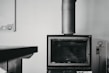 A black and white image featuring a modern, rectangular fireplace with a large metal chimney extending upwards. On the right side, there is a set of fireplace tools, including a poker, tongs, and a brush, hanging from a stand. A table is partially visible on the left side of the image. The background is a plain, light-colored wall with a small, partially visible item in the upper left corner, possibly a calendar or bulletin board.
