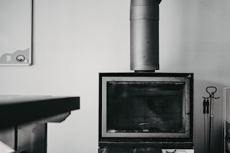 A black and white image featuring a modern, rectangular fireplace with a large metal chimney extending upwards. On the right side, there is a set of fireplace tools, including a poker, tongs, and a brush, hanging from a stand. A table is partially visible on the left side of the image. The background is a plain, light-colored wall with a small, partially visible item in the upper left corner, possibly a calendar or bulletin board.