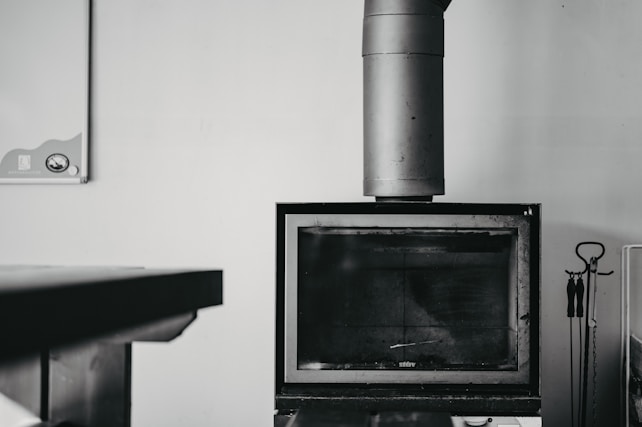 A black and white image featuring a modern, rectangular fireplace with a large metal chimney extending upwards. On the right side, there is a set of fireplace tools, including a poker, tongs, and a brush, hanging from a stand. A table is partially visible on the left side of the image. The background is a plain, light-colored wall with a small, partially visible item in the upper left corner, possibly a calendar or bulletin board.