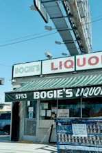 A liquor store named Bogie's Liquor located at 5753. The storefront has a striped green and white awning with the store's name written on it. Above it is a large rectangular sign with 'BOGIE'S' in green letters and 'LIQUOR' in red letters. There is an outdoor bulletin board with various postings, including one that says 'THE WORLD IS WAITING FOR YOU'. A large billboard structure is visible above the store.