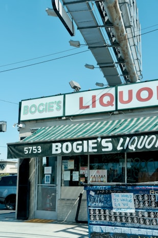 A liquor store named Bogie's Liquor located at 5753. The storefront has a striped green and white awning with the store's name written on it. Above it is a large rectangular sign with 'BOGIE'S' in green letters and 'LIQUOR' in red letters. There is an outdoor bulletin board with various postings, including one that says 'THE WORLD IS WAITING FOR YOU'. A large billboard structure is visible above the store.